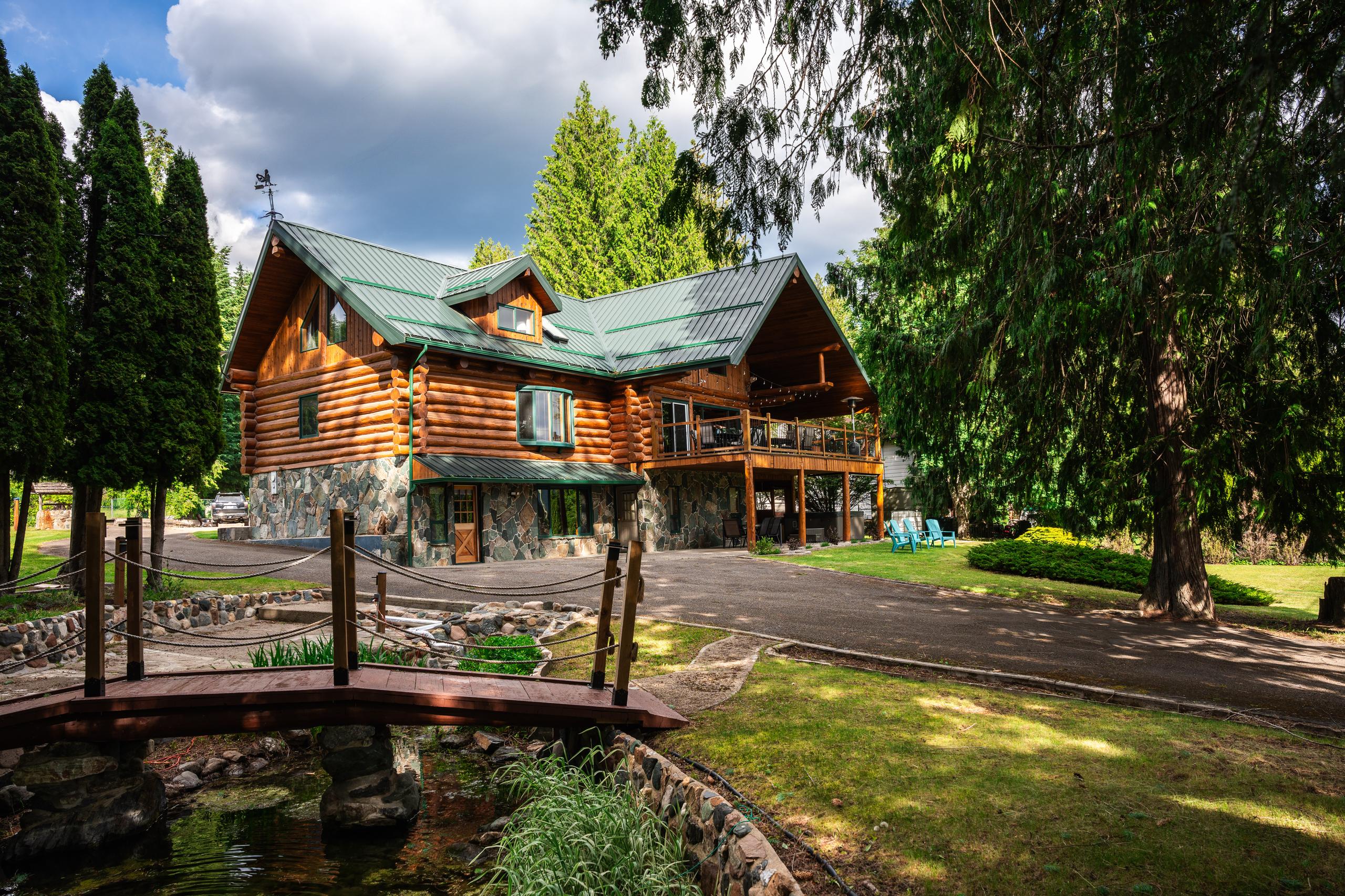 The log home in summer, its green metal roof above a stone base, with the footbridge over the garden pond in the foreground.