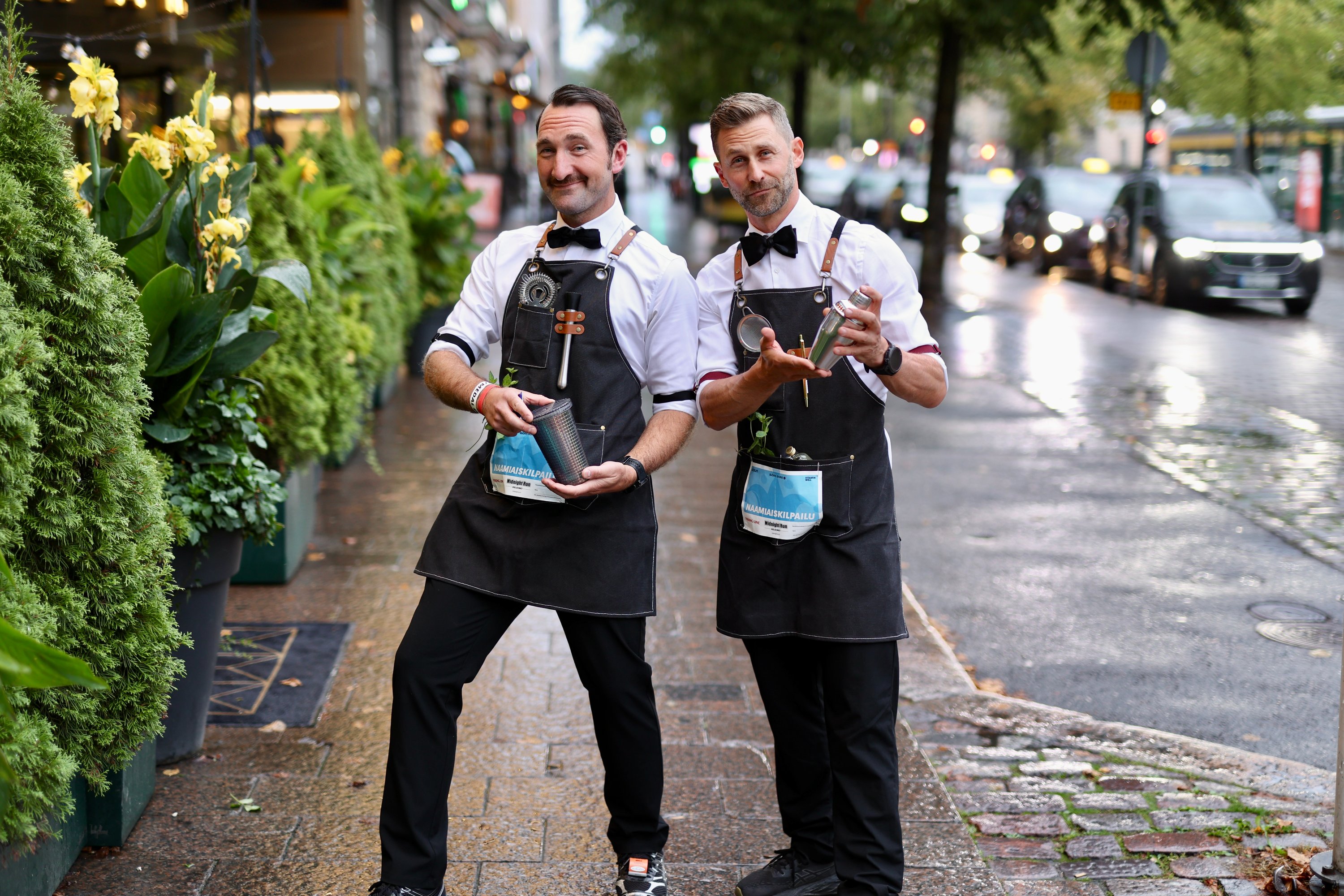 Simon och Fredrik i förkläde och fluga med varsin shaker på en regnig gata i Helsingfors