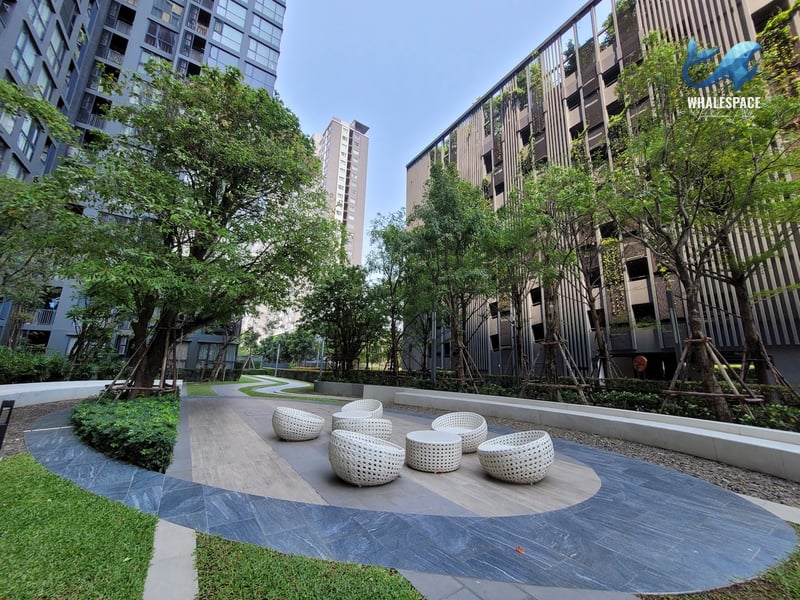 Lush communal garden area featuring modern circular seating arrangements amidst manicured greenery, with tall residential buildings in the background.