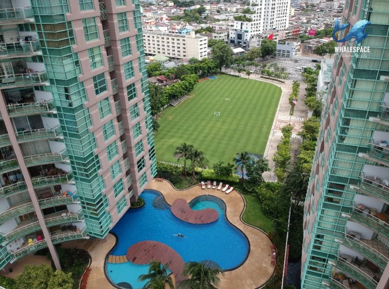 Aerial view of a uniquely shaped outdoor swimming pool surrounded by tropical landscaping, adjacent to a large manicured green field and residential towers.