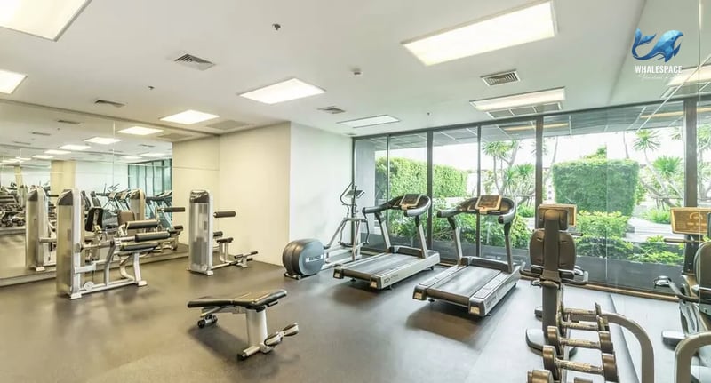 Spacious and bright fitness center equipped with various cardio and weight machines, featuring large windows overlooking a green garden.