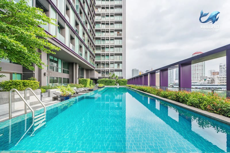 Outdoor swimming pool with shimmering blue water, surrounded by lush greenery and modern building architecture, featuring city skyline views.