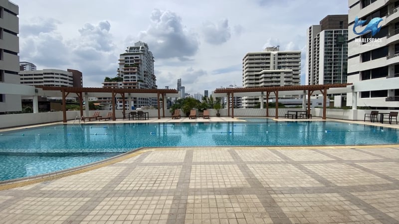Expansive swimming pool with a tiled deck and shaded lounge areas, set against a backdrop of city skyscrapers.