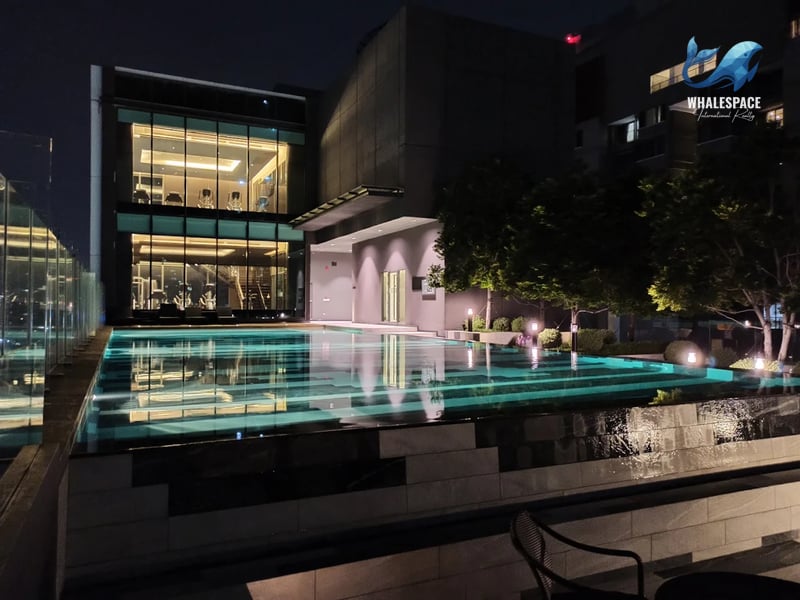 Outdoor swimming pool illuminated at night, with a modern building featuring a visible fitness center through large glass windows in the background.