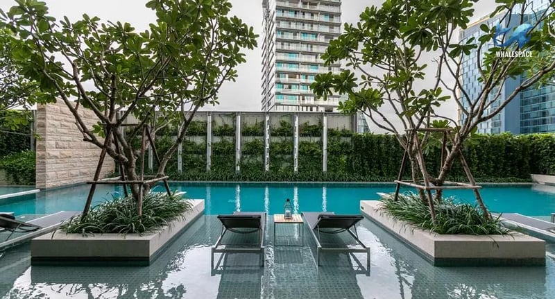 Resort-style swimming pool surrounded by lush trees and modern lounge chairs, with a high-rise building in the background
