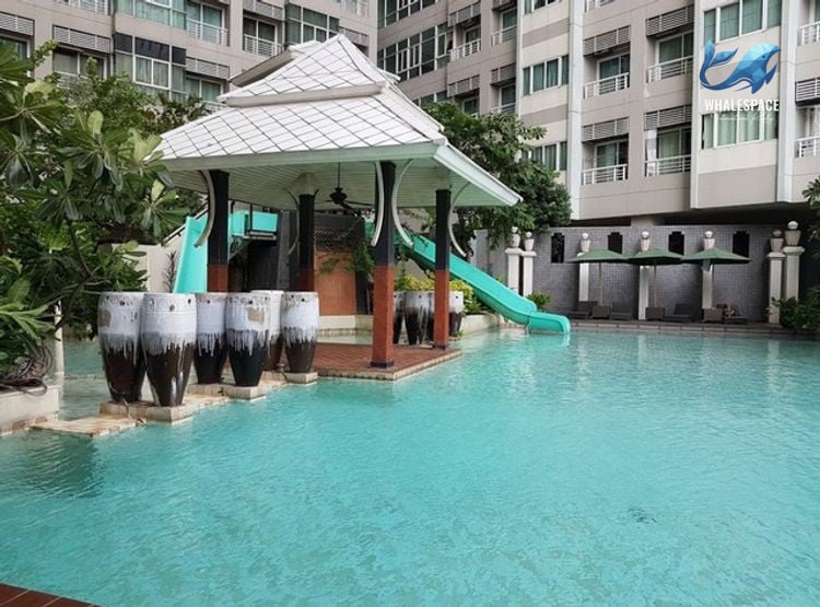 Resort-style swimming pool with a shaded cabana and water slide, surrounded by lush greenery and apartment buildings.