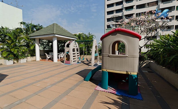 Children's playground with slides and swings on a tiled surface, shaded by a green-roofed gazebo with seating.