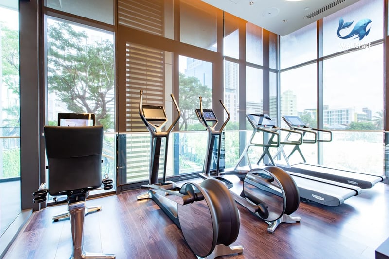 Well-equipped fitness room with various exercise machines, wooden floors, and large windows providing natural light and exterior views.