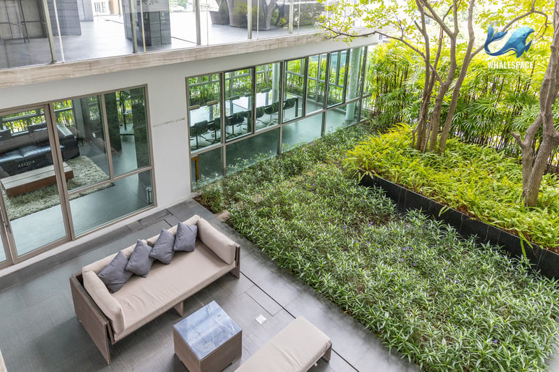 Comfortable lounge area with a beige sofa and coffee table, adjacent to a vibrant green garden and meeting rooms with large glass windows.