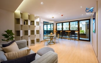 Spacious communal lounge area with light wood flooring, a grey sofa, small round tables, and bar seating by large windows.