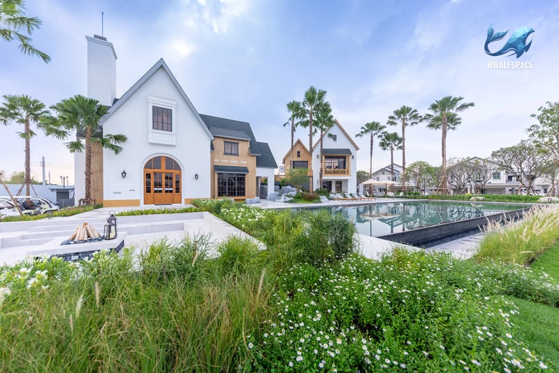 Elegant white house with a distinctive gabled roof and a grand arched wooden entrance, set against a backdrop of lush greenery and palm trees beside a serene swimming pool.