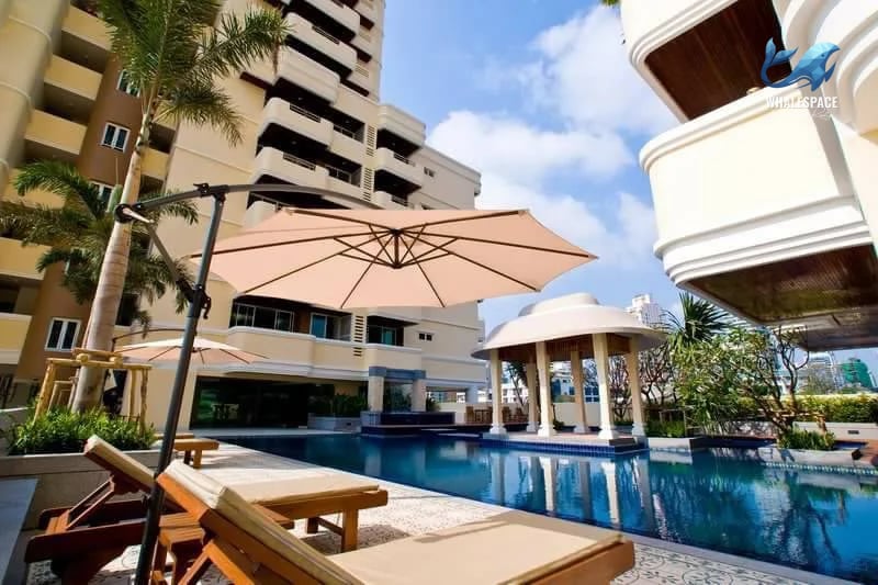 Resort-style swimming pool area with lounge chairs and a shaded cabana, surrounded by lush palm trees and modern architecture.