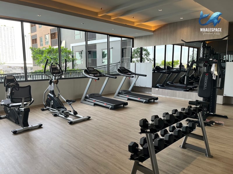 Well-equipped fitness center with various cardio machines and a dumbbell rack, set against large windows offering views of the surrounding area.