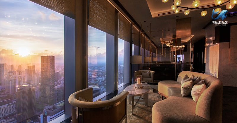 Elegant lounge area with floor-to-ceiling windows offering panoramic city views at sunset, furnished with contemporary seating and a unique chandelier.