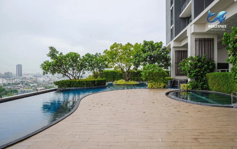 Infinity edge swimming pool with lush trees and a panoramic city skyline view under an overcast sky.