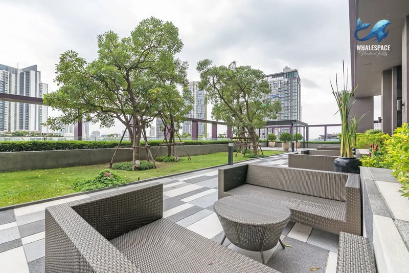 Outdoor lounge area with contemporary woven seating and tables, set amidst manicured trees and paved sections, with city buildings in the background.