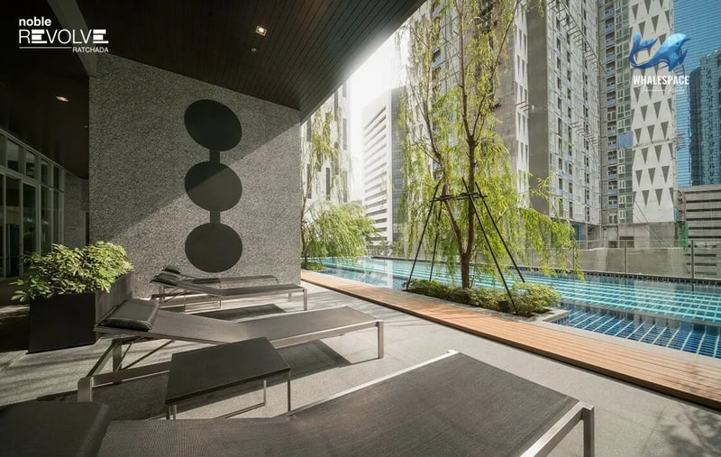 Relaxing outdoor swimming pool area with sun loungers and lush greenery against a city backdrop.