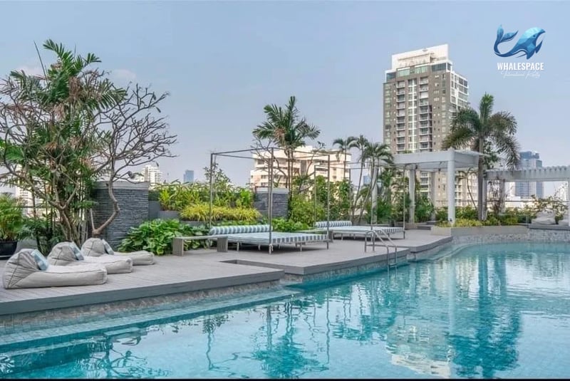 Outdoor swimming pool area with sun loungers, daybeds, lush plants, and a backdrop of city buildings under a clear sky.
