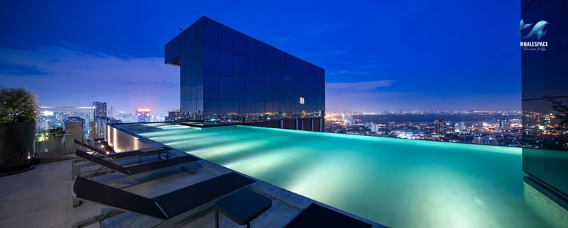 Infinity swimming pool overlooking a vibrant city skyline at dusk, with lounge chairs on the deck.