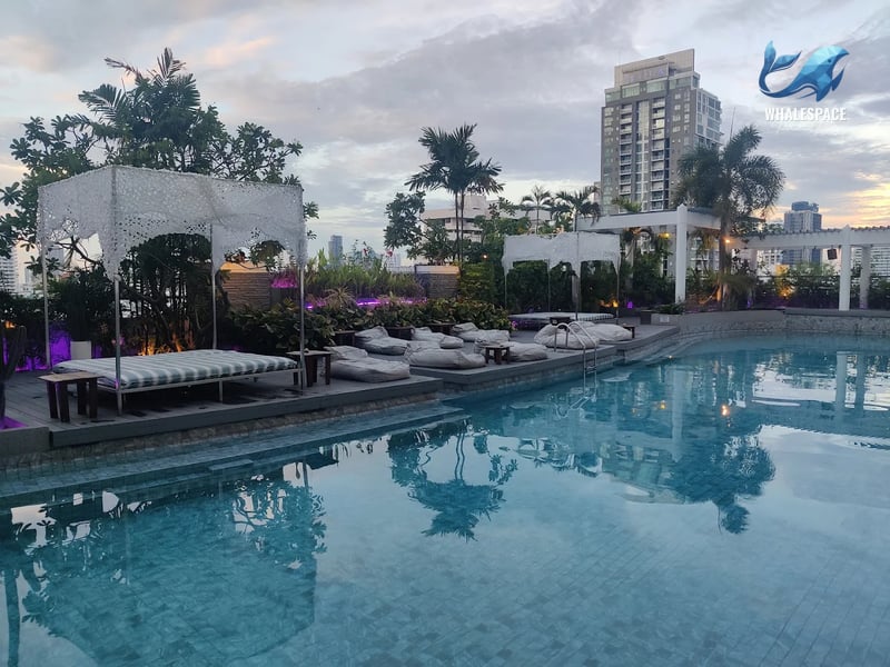 Swimming pool area at sunset, featuring elegant daybeds with canopies, comfortable bean bags, and city views.