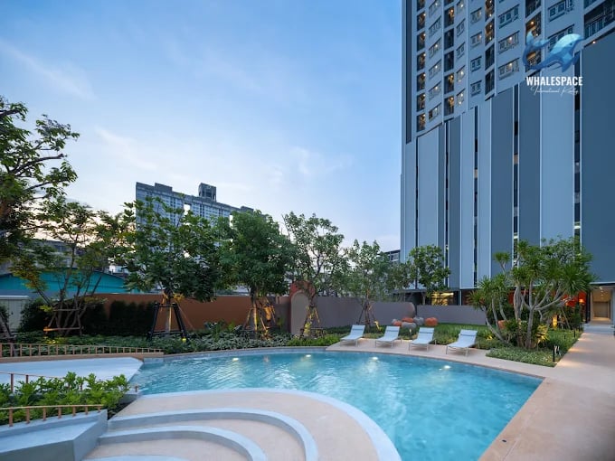 Resort-style swimming pool surrounded by lush greenery and lounge chairs, with a modern high-rise building in the background.