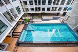 Overhead view of an outdoor swimming pool with blue mosaic tiles, surrounded by a wooden deck and modern condominium buildings.