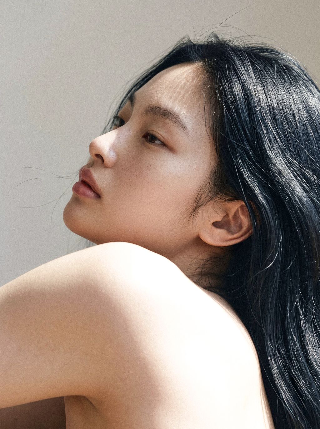 Profile of a young Asian woman with long dark hair and bare shoulders, looking up in soft natural light.