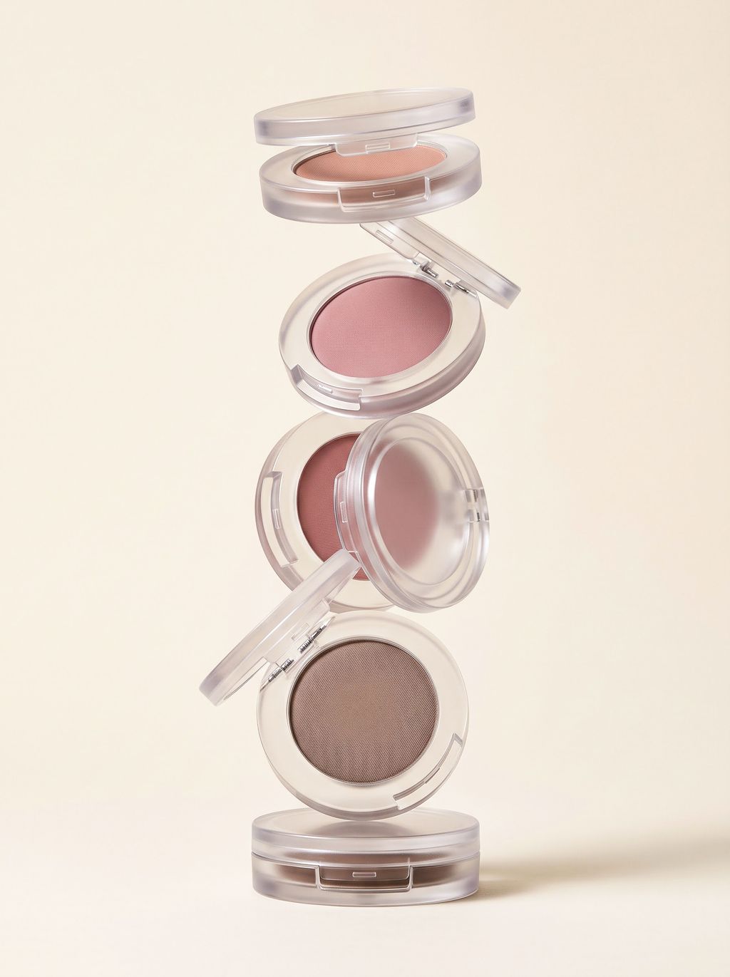 Stack of five round makeup compacts with nude, pink, rose, and brown shades on a beige background.