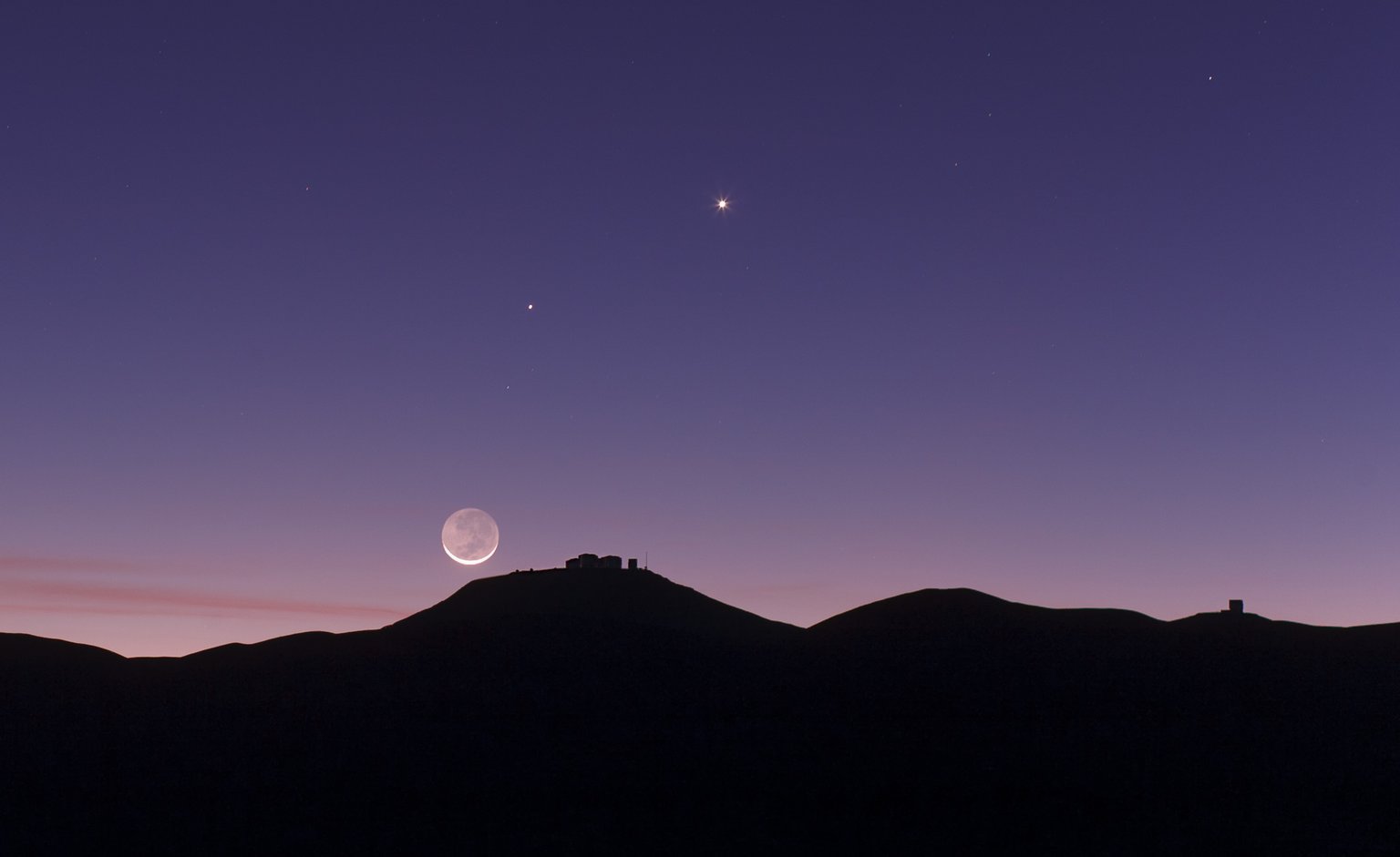 Srpek Měsíce s popelavým svitem nad siluetou observatoře Paranal v Chile.