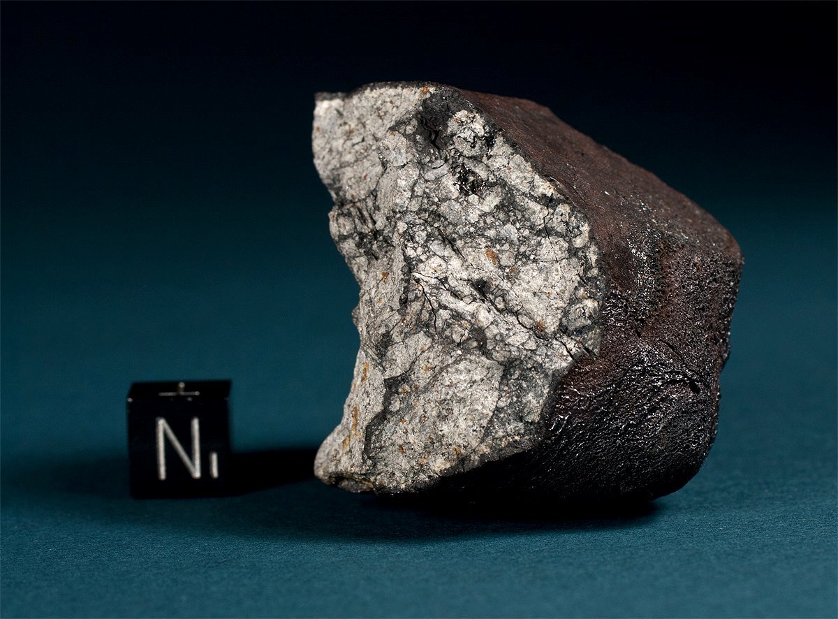 Úlomek čeljabinského meteoritu s tmavou kůrou a světlým lomem vedle centimetrové kostky.