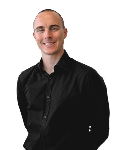 Adam Barbarich, with short hair and a black button-up shirt, stands against a plain, light background, smiling with hands behind their back.