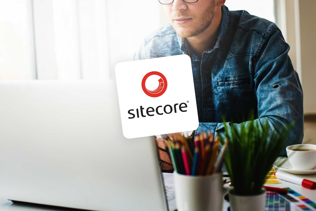 A man wearing glasses and a denim shirt works on a laptop at a desk with colourful pencils and pens. The Sitecore logo is prominently displayed in the centre, highlighting his role as a Sitecore Developer.