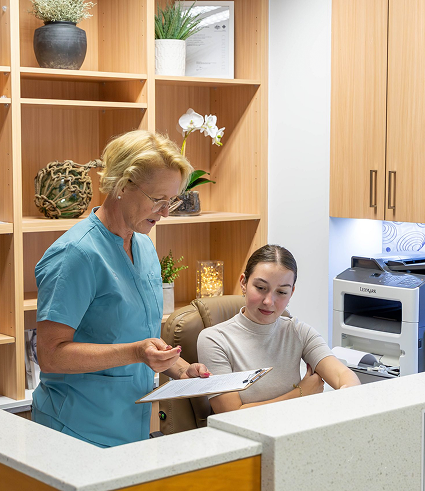 A healthcare professional in blue scrubs is showing a clipboard to a seated woman at the Perth City Dental reception desk in a modern office with wooden shelving and office equipment in the background.