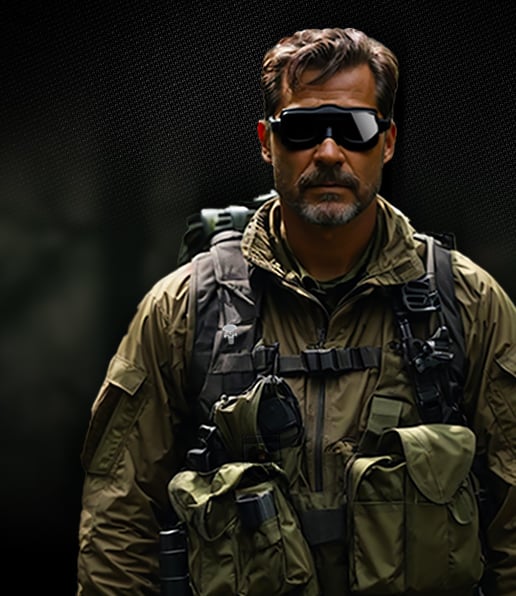 A man in tactical military gear with sunglasses and a beard stands against a dark, blurred background. He wears an olive green jacket, a tactical vest with multiple pouches, and carries a rucksack.