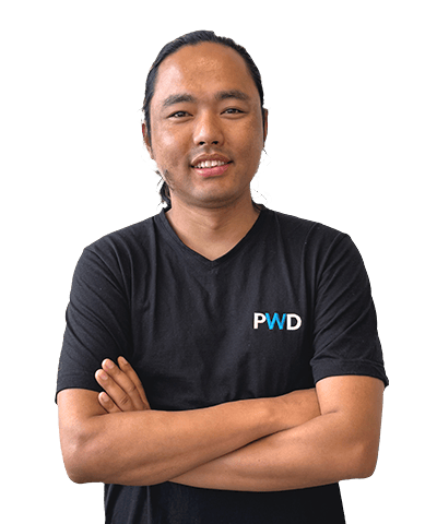 A person with long hair tied back is standing with arms crossed, wearing a black T-shirt with the letters PWD on it, and smiling at the camera against a plain white background.