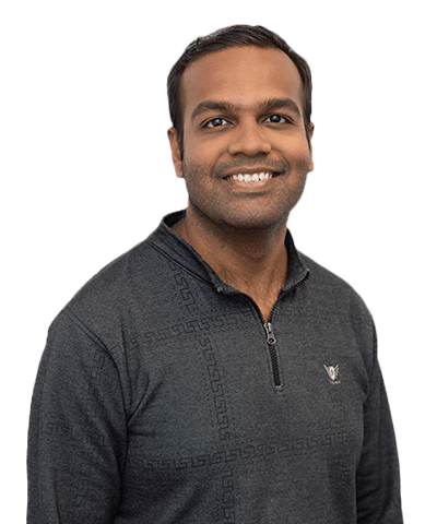A man with short dark hair smiles at the camera. He is wearing a dark, patterned quarter-zip jumper and is posed against a plain, light background.