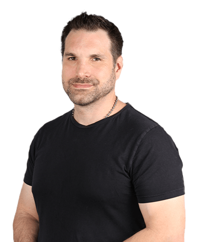 Chris Messina, a man with short dark hair and a beard, wearing a black T-shirt and a chain necklace, stands against a plain light background, smiling slightly at the camera.