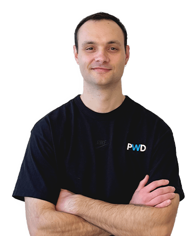 Tristram Vickers, a man with short dark hair, wearing a black T-shirt with PWD on it, stands with his arms folded and smiles slightly against a plain white background.