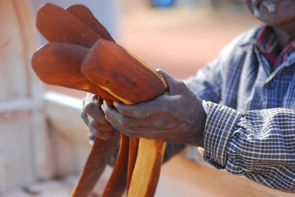 A person wearing a checked shirt holds several reddish-brown wooden boomerangs, displaying them with both hands in an outdoor setting. These unique pieces of Indigenous art are crafted with skill and tradition. Their face is partially out of shot.