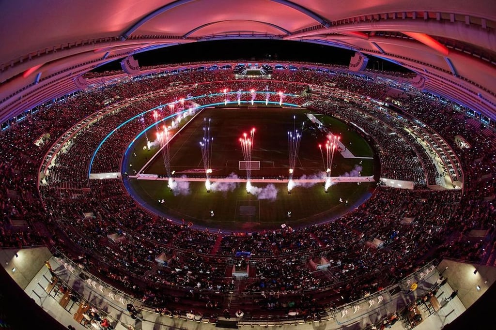 A large stadium filled with spectators under bright lights, with colourful fireworks displays and smoke on the football pitch during an event or celebration.