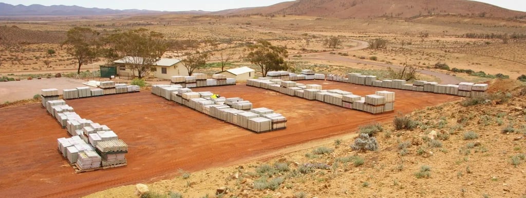 Aerial view of stacks of building materials arranged on a red dirt lot in a remote, arid landscape with small buildings and sparse vegetation. Hills and a winding road are visible in the background.