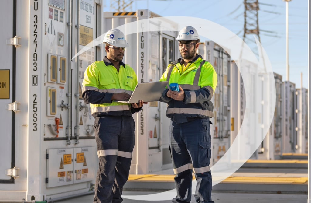 Two workers in high-visibility uniforms and helmets stand outside at an industrial facility, looking at a laptop. Shipping containers with control panels line the area. It’s sunny, power lines are visible, and the site reflects Partum Engineering’s expertise.