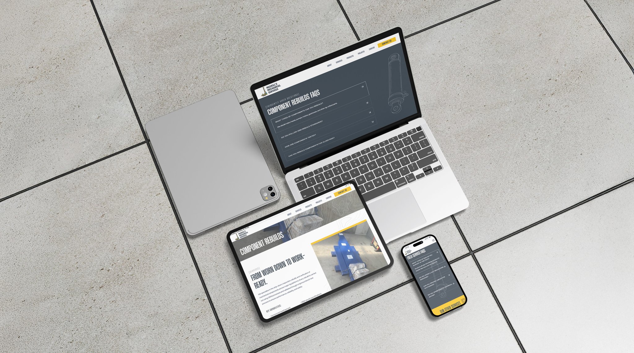 A laptop, tablet, and two mobile phones are displayed on a light grey tiled floor. All devices show the Maskills Mechanical Services website with a dark and yellow colour scheme. One mobile phone and the laptop are closed.