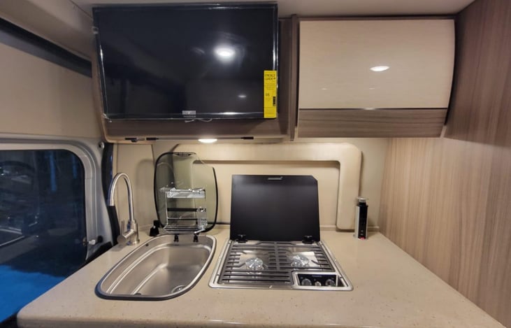 The kitchen is equipped with a sink, a two-burner stove, and a microwave, and there is a 24" TV mounted above. The TV swings out to more storage.