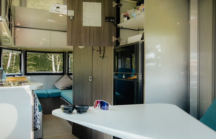 Front to back view showing open storage above fridge, and huge windows in back. All windows have black-out blinds for sleeping and they also have screens and can open wide for plenty of fresh air.