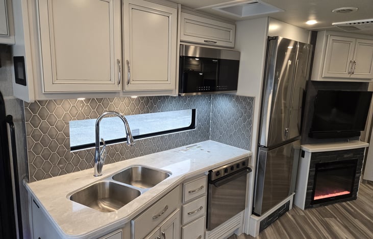 View of RV galley area with double sinks, convectional microwave, three burner stove area and oven, electric refrigerator/freezer, electric fireplace, and LED TV screen with sound bar.
