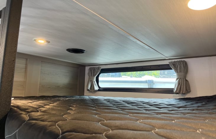 Loft bunk has window with curtains and A/C vent. Light switch, electrical outlet, and small in-wall open storage  at end of bunk