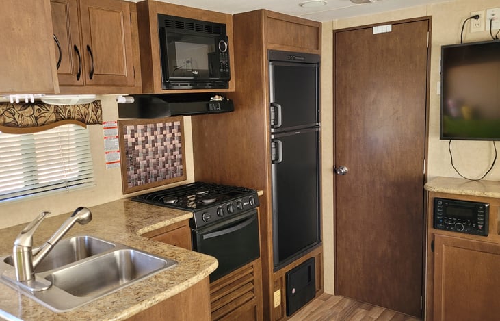 2-vessel Kitchen Sink, Countertop, Oven & Range, Microwave, Refrigerator & Freezer, Bathroom Door, TV, Cabinets overhead containing dishes, cups, etc.