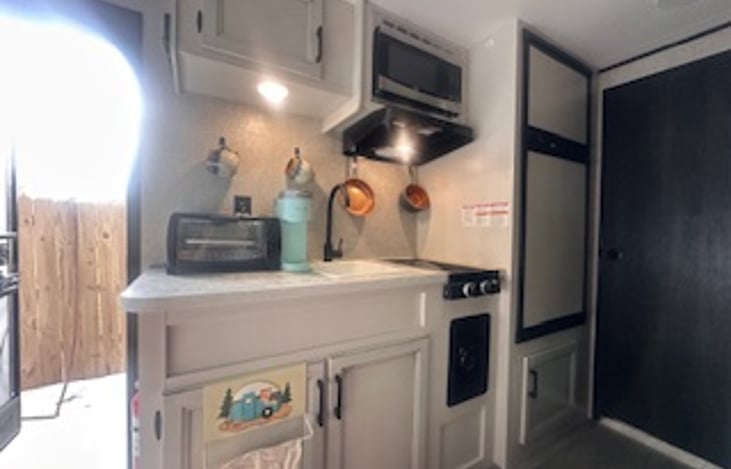 Kitchen area houses small refrigerator, freezer, two eye propane stove, microwave, kuerig, toaster oven, pots, pans, cups, plates, bowls, and utensils,