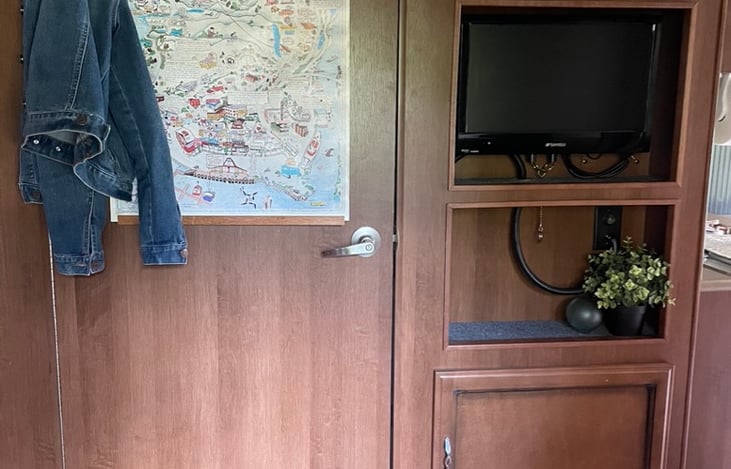 The hallway and bathroom door, facing the bunk beds/small dinette. There is a tv located here and storage above and below.